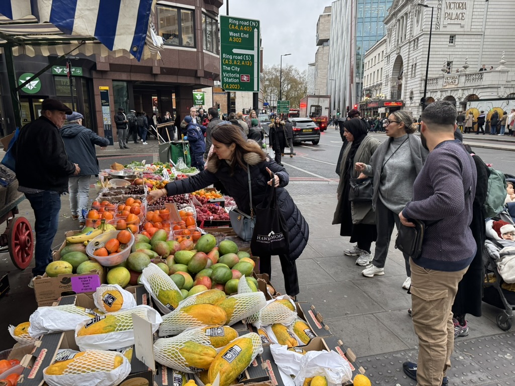 Shoppers and passersby inspect Mick's wares which include several kinds of tropical fruit and bulk quantities of berries.